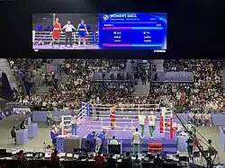 Khelif e Carini estão no ringue de boxe na segunda eliminatória, a aguardar o anúncio da vencedora. Khelid levanta o punho triunfante, enquanto Carini olha para o chão.