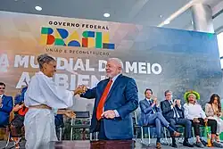 Lula e Marina Silva cumprimentam-se, em pé, em cerimônia no Palácio do Planato. Atrás deles, aparecem sentadas algumas autoridades, como o ministro Fernando Haddad, e personalidades da sociedade civil, como o cacique Raoni. Ao fundo, há um painel com a frase "Dia Mundial do Meio Ambiente" e a logomarca do terceiro mandato do governo Lula.