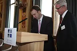 Dois homens brancos e de terno observam algo em um púlpito na esquerda da imagem