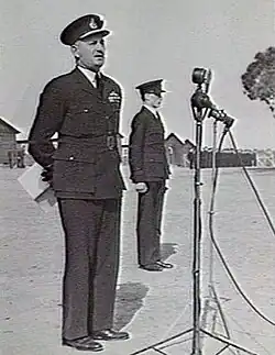 Man in dark military uniform with peaked cap in front of microphone