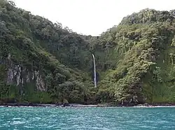 Vista de uma cachoeira alta e estreita descendo por uma encosta coberta de vegetação densa na Isla del Coco, Costa Rica. A cachoeira deságua próxima à costa, com o mar azul-turquesa em primeiro plano.
