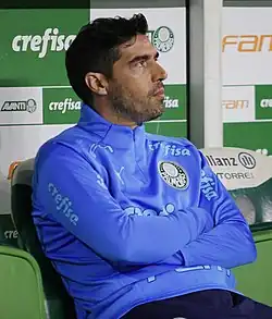 Um homem sentado num banco, olhando para cima, com os braços cruzados.