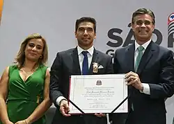 Uma mulher, ao lado de dois homens, que estão segurando um quadro com uma placa. Todos estão com feições felizes.