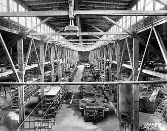 Vista interior de um grande galpão industrial com asas de aeronaves penduradas e equipamentos.