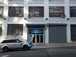 Faixada do prédio da American Musical and Dramatic Academy mostrando a porta frontal, algumas janelas e um carro estacionado a esquerda.