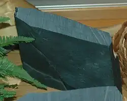 Amostra bruta de argilita, com textura compacta, bordas irregulares e cor cinza-escura.