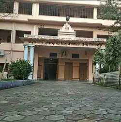 Entrada principal de uma escola. Na fachada é possível ver o nome da escola escrito em inglês e em Bengali.