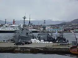 Imagem do porto da base naval mostrando 5 navios militares em primeiro plano e 2 navios maiores ao fundo, na distância além do porto se encontra uma cidade e um morro a direita.