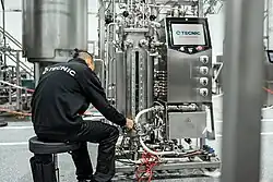 Um técnico ajusta uma válvula num grande biorreator de aço inoxidável ligado a um painel de controlo.