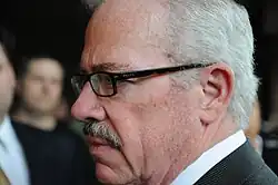 Close do lado esquerdo de um homem de cabelos grisalhos com bigode, usando óculos Prada de armação grossa