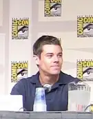 Brian J. Smith, um homem branco, com cabelo castanho escuro curto, em uma mesa de discussão com um microfone em sua frente.