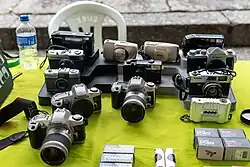 Coleção de câmeras fotográficas vintage e digitais expostas em uma Feira de Antiguidades da Praça XV. O conjunto inclui diversos modelos como Canon EOS, Nikon, Yashica e Minolta, algumas equipadas com lentes zoom. Também estão expostas câmeras compactas em tons bege e prata, junto com várias caixas do filme fotográfico Fomapan e outros filmes dispostos na parte inferior do display.