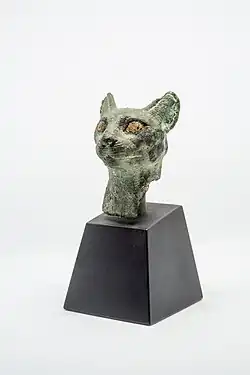 Cabeça de uma estatueta representando um gato com os olhos e narinas dourados (ouro) e as orelhas furadas para fixação de brincos. Incisões definem a boca e os bigodes. As órbitas oculares foram cobertas ou preenchidas possivelmente com ouro. A figura está fragmentada abaixo do pescoço mostrando o interior que não apresenta sinais de substância de preenchimento para fundição. A extremidade da orelha direita está quebrada.