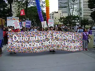 Grupo de mulheres marchando pela calçada segurando uma faixa com os dizeres “Outro mundo é possível sem racismo, machismo e lesbofobia - Marcha Mundial das Mulheres
