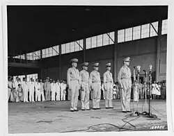 MacArthur stands in uniform at four microphones on stands. Behind him four men in army uniforms stand at attention. There are viewed by a large crowd of well-dressed men, women and children in skirts, suits and uniforms.
