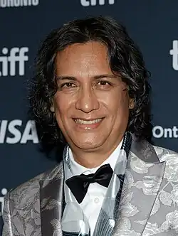 Uma foto de Carlos Diehz usando um terno prateado e uma gravata borboleta preta torta.