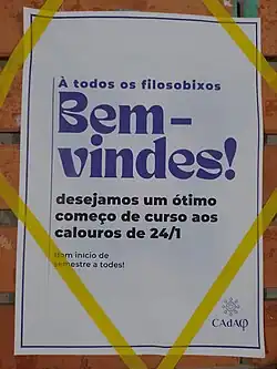 Cartaz com fundo creme e letras rochas com o texto "Bem-Vindes - desejamos um ótimo começo de curso aos calouros de 24/1"