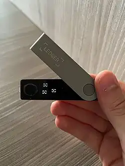Mão segurando uma carteira hardware Ledger Nano X sobre fundo de madeira