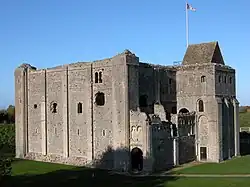 Um prédio quadrado de pedra cinza com fendas verticais estreitas no primeiro andar e janelas mais largas no segundo. O topo do castelo parece podre e não há telhado, exceto sobre uma torre anexada à torre.