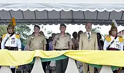 Aldo aparece em posição de sentido num estande, de terno, junto de dois outros comandantes militares e cercados por dois soldados em uniforme tradicional