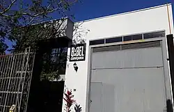 Fachada da Cervejaria Babel, nas cores branca e cinza, com portão alto à direita, vegetação à esquerda e letreiro com a logomarca ao centro.