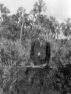 Fotografia em preto e branco de um homem seminole vestindo traje tradicional com colete, segurando um rifle, de pé entre palmeiras, olhando para o observador