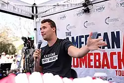 Charlie Kirk em frente a um painel com logotipos da Turning Point USA e "American Comeback Tour" escrito em fonte grande. Sua camisa diz "ANTI-DEI PRO-MERIT"