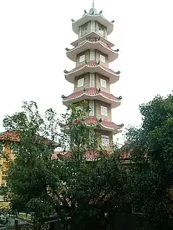 Uma torre de sino octogonal com sete níveis, azulejos ornamentados e cercada por plantas.