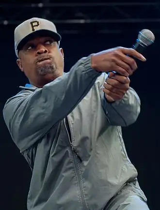 Rapper Chuck D fazendo o movimento de um taco de baseball com o microfone enquanto performa. Ele usa um boné do time de baseball Pittsburgh Pirates.