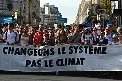 Grupo de pessoas diversas vistas de frente e segurando um cartaz à luz do dia em uma avenida de Paris