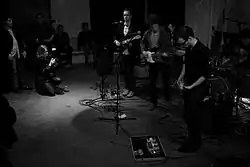 Uma banda vestida com camisas pretas tocando música, cercada por pessoas