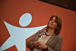 Catarina Martins em frente a parede vermelha com o símbolo branco do Bloco de Esquerda. Catarina tem o cabelo pelos ombros, tem um colar azul, uma blusa axul e um blazer castanho.