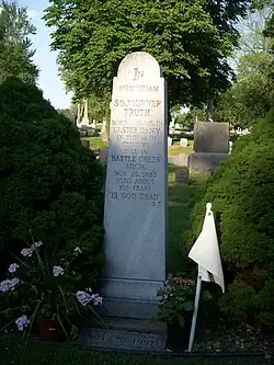Foto de um túmulo com dois vasos de flores e uma bandeira branca ao seu lado. Ao seu redor, há árvores e outros túmulos. Em seu epitáfio, traduzindo de inglês para português, está escrito: Em memória. Sojourner Truth. Nasceu escravizada no Condado de Ulster, Nova Iorque no século 18. Morreu em Battle Creek, Michigan, em 26 de novembro de 1883. Com idade aproximada de 105 anos. "Deus está morto".