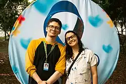Foto dos participantes João Victor Ramos e Brendaly Januário. Eles estão num parque, à frente de um cartaz com um símbolo de laço de fita representando o autismo. Ele está vestido com uma blusa amarela e preta, com um cordão verde com símbolo do girassol. Ele é branco, tem cabelos castanhos, olhos verdes, óculos e barba no queixo. Brendaly está vestida com uma blusa branca com desenhos azuis. Ela é branca, tem cabelos pretos cacheados e usa óculos.