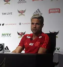 Um homem sentado em uma cadeira, vestindo uma camisa vermelha, com uma mesa à sua frente, num ambiente com pouca iluminação. Ele está olhando para atrás da câmera, sem expressão definida