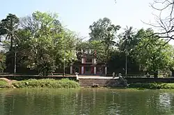 Um lago verde diante da entrada do templo; escadaria de pedra até um portão vermelho de dois níveis; área cercada por arbustos e árvores.