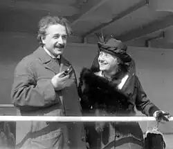 Albert Einstein, parecendo relaxado e segurando um cachimbo, está ao lado de uma sorridente e bem-vestida Elsa, que usa um chapéu chique e um casaco de pele. Ela está olhando para ele.