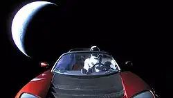 Photograph of the front of a red convertible sports car floating in space. There is a humanoid figure in the driving seat. In the background, partially illuminated in a crescent shape, is planet Earth.