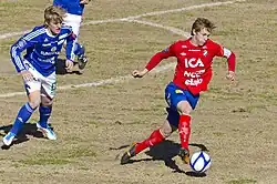 jogador Emil Krafth vestido de vermelho conduzindo a bola com o pé esquerdo em um campo de futebol enquanto existem jogadores de azul correndo atrás.