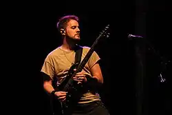 Eric tocando guitarra e olhando para o seu lado esquerdo ante um fundo negro