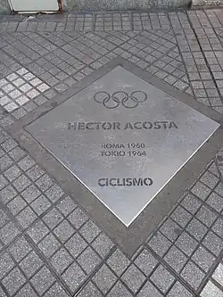 Fotografia de uma placa quadrada ou em forma de pipa prateada embutida na calçada com o nome do ciclista e as duas competições olímpicas em que participou, nomeadamente Roma 1960 e Tóquio 1964