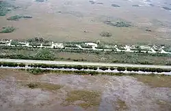 Fotografia colorida tirada do ar mostrando os Everglades divididos por uma autoestrada; na parte inferior está um campo de erva-serrada inundado com água ladeado por um canal cheio; na parte superior estão algumas casas e um campo de erva-serrada seco
