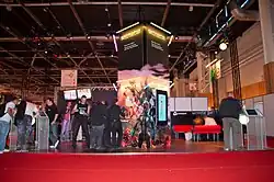 Imagem de uma feira onde se vê o estande do jogo e pessoas interagindo com o estande.