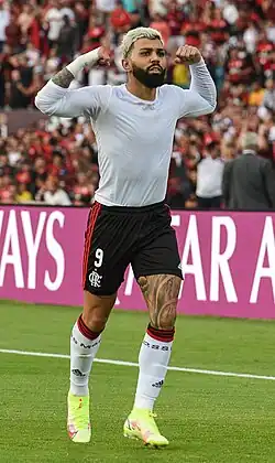 Um homem com a pele morena, com os cabelos loiros e barba morena cheia correndo lentamente. Ele está com os seus dois braços na altura do rosto, flexionando os bíceps e com a cara séria.