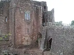 Um castelo, com uma torre de frente plana voltada para o espectador com uma janela de vitral no meio dela; uma calçada de pedra está à direita da imagem, levando a um portão à direita da torre – um arco parcialmente preenchido está apoiando a calçada.