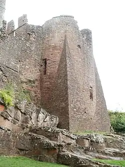 Uma enorme torre de castelo, sentada no topo de um afloramento rochoso acidentado; um enorme esporão de pedra angular se projeta da base da torre em direção ao espectador.