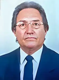 Retrato formal do ex-governador José Muniz Ramos. Ele é um homem de meia-idade, com pele clara, cabelos curtos e escuros, levemente grisalhos, penteados para trás. Usa óculos de armação fina e tem expressão séria. Está vestido com terno escuro, camisa branca e gravata azul-marinho, em um fundo claro e neutro.