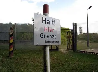 Um aviso em branco em um poste com a inscrição em alemão "Halt! Hier Grenze" (Pare! Fronteira aqui) e abaixo, em letra menor, "Bundesgrenzschutz" (Guarda da Fronteira Federal). No fundo, uma cerca de fios metálicos com uma porta aberta, e atrás dela estão árvores e uma torre de vigilância.