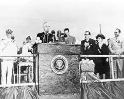 Fotografia a preto e branco do Presidente Harry Truman de pé num púlpito com o selo presidencial num palco com pessoas atrás dele a aplaudir