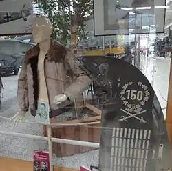 Foto colorida de uma vitrine, dentro com um boneco manequim vestindo uma jaqueta de pele ao lado de um leme de cauda de avião. O leme apresenta várias marcações, no topo o número 150, rodeado por uma coroa de flores. Abaixo da coroa há duas espadas cruzadas e numerosas listras brancas.